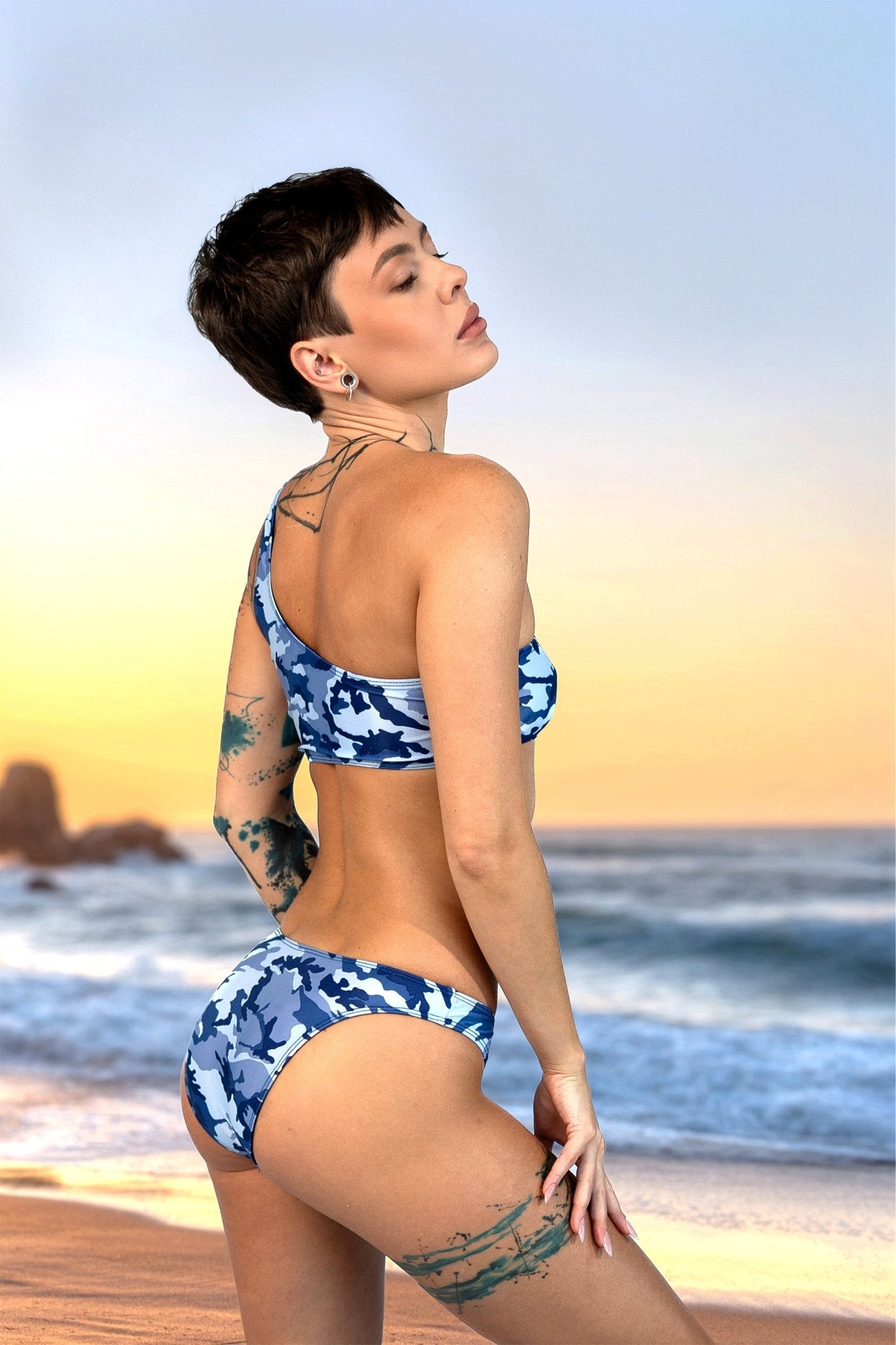A woman with short hair and tattoos poses on a beach at sunset, wearing Brigitewear International's Camo Cannes Ocean Wave Asymmetrical Top.
