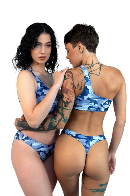 Two women wear Brigitewear’s Camo Cannes Ocean Wave Asymmetrical Top swimsuits on the beach—one faces forward, the other has her back to the camera.