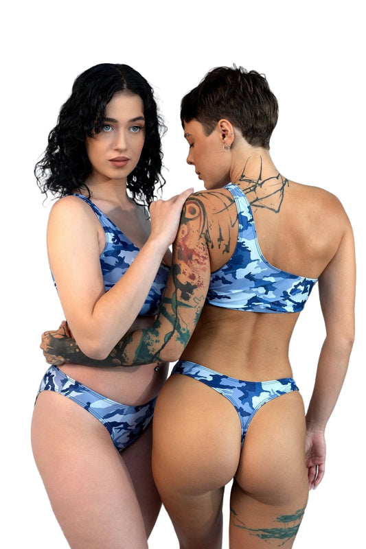 Two women wear Brigitewear’s Camo Cannes Ocean Wave Asymmetrical Top swimsuits on the beach—one faces forward, the other has her back to the camera.