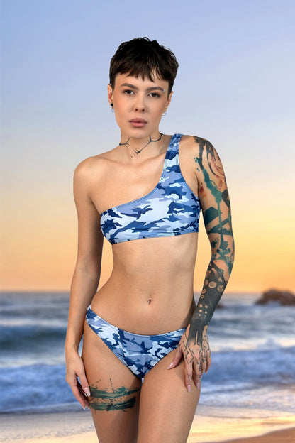 A woman with short hair and tattoos wears the Brigitewear Camo Cannes Ocean Wave Asymmetrical Top by Brigitewear International, LLC, standing on a beach at sunset.