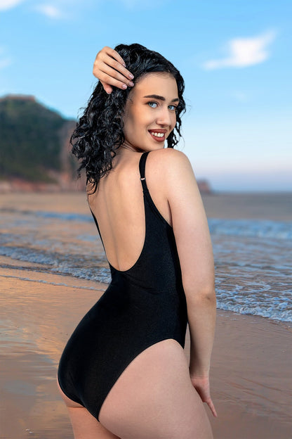 A woman smiles on a sandy beach with ocean and cliffs behind her, wearing the Ring Me Up One-Piece by Brigitewear International, LLC.