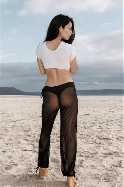 On a sandy beach, a woman faces away wearing Brigitewear’s Sheer Mesh Pants—Beach Cover-Up Trousers by Brigitewear International, LLC, styled with a white crop top.