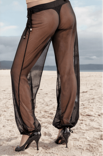 A person wearing Brigitewear Sheer Mesh Pants – Beach Cover-Up Trousers by Brigitewear International, LLC and high heels stands on a sandy beach, facing away from the camera.