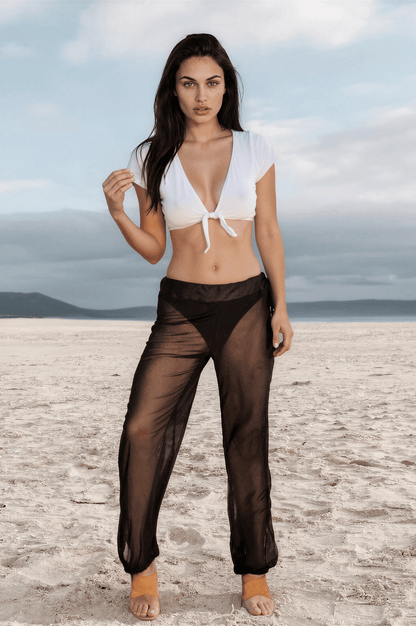 A woman stands on a sandy beach beneath a cloudy sky, wearing Brigitewear International, LLC’s Sheer Mesh Pants - Beach Cover-Up Trousers paired with a white crop top.