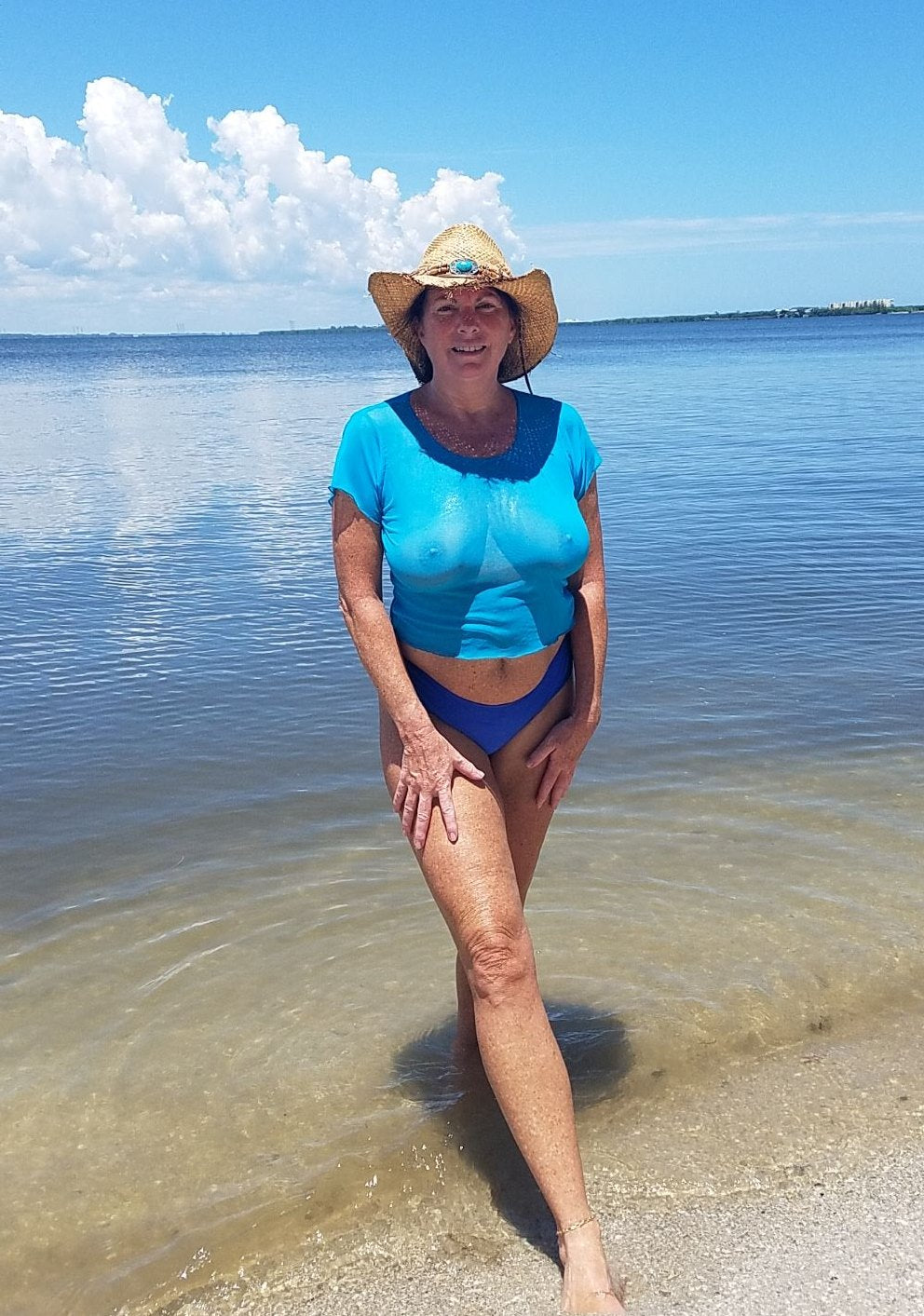 A woman in Brigitewear beachwear and a sunhat stands in shallow water at a sunny beach, enjoying the clear sky.