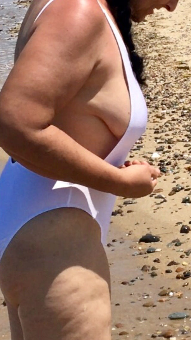A person in a white swimsuit stands on a rocky beach, looking down and holding something in their hand.