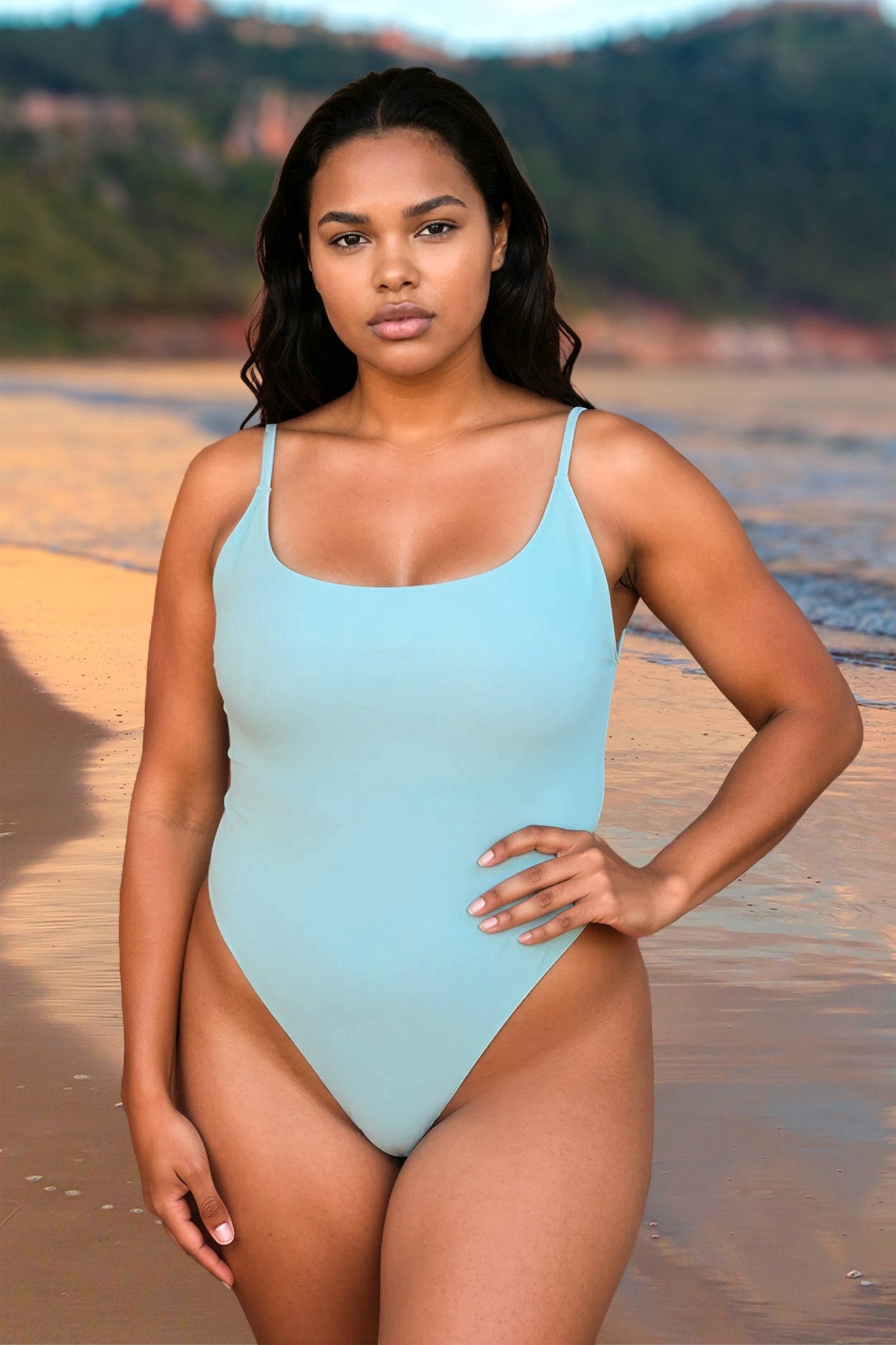 A woman wears the St. Tropez One Piece Thong Swimsuit by Brigitewear International, LLC, standing on a sandy beach with water and cliffs in the background.