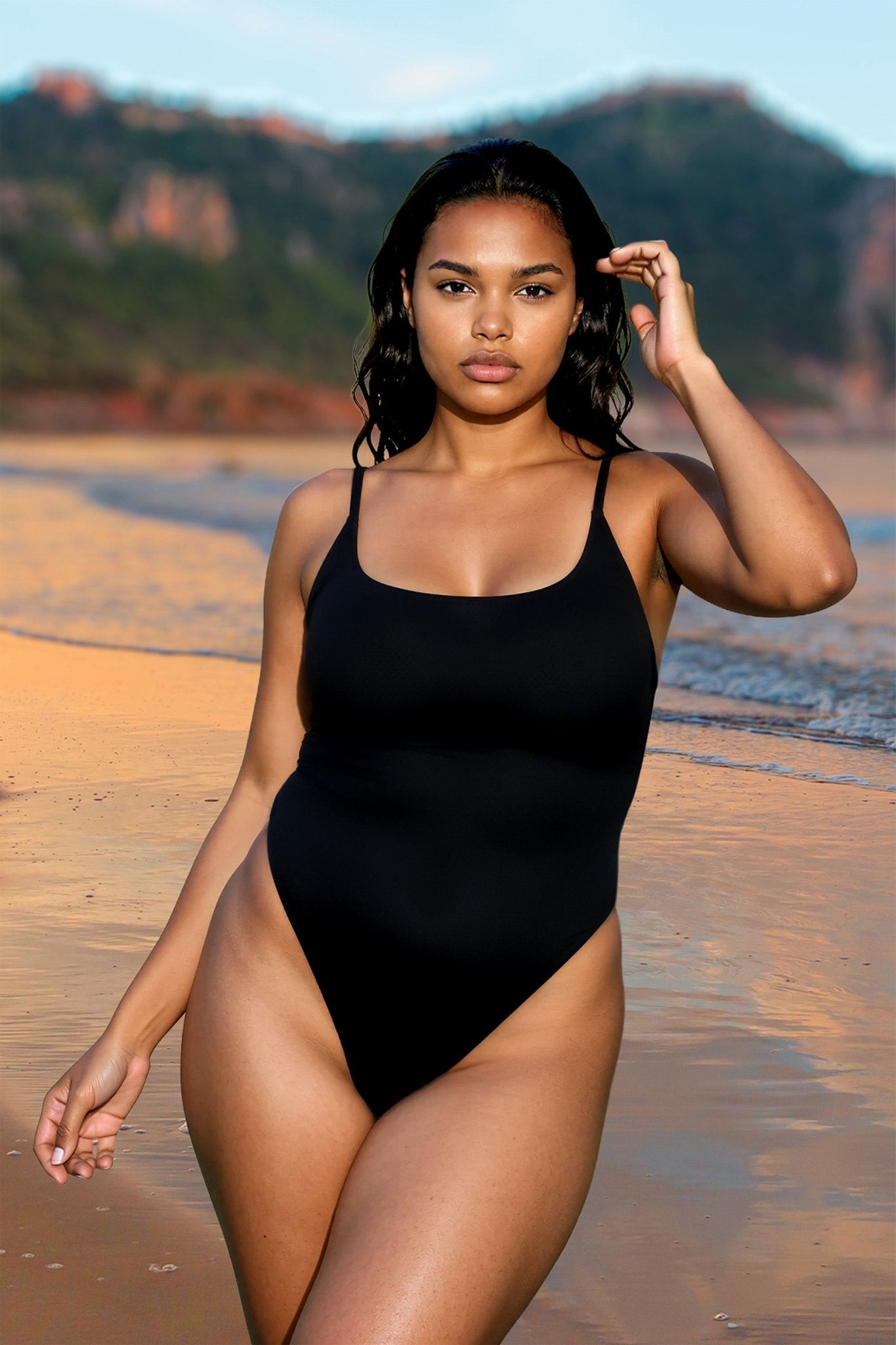 Woman wearing the Brigitewear St. Tropez One Piece Thong Swimsuit—Sexy High-Cut Sheer Swimwear—stands on a beach with mountains and water in the background.