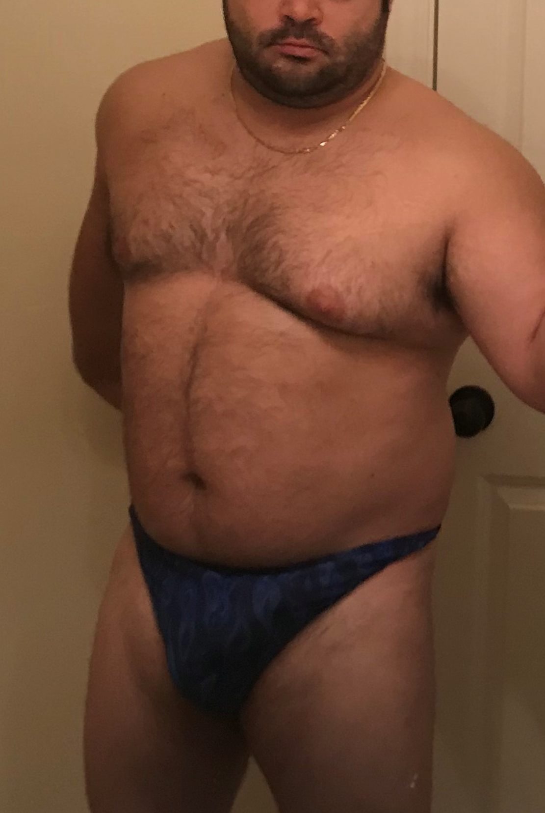 A man wearing a blue thong stands in front of a door, showing his chest and torso.