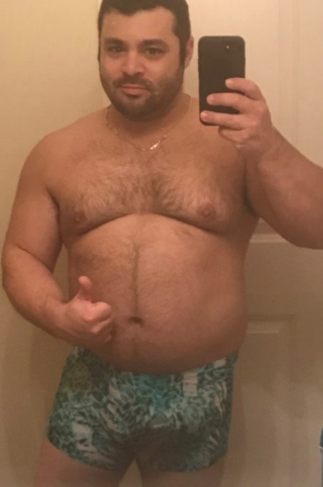 A man in patterned shorts, perfect for resort wear, takes a mirror selfie and gives a thumbs up in a bathroom.