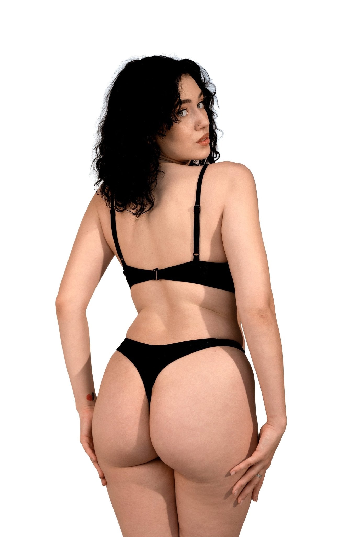 Woman with curly dark hair models the Tri-Ring Sexy Thong by Brigitewear International, LLC, facing away and looking over her shoulder against a white background.