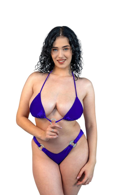A woman with curly dark hair smiles in a Brigitewear International V-Bottom Thong & Rio Bikini Bottom, standing against a plain light gray background.