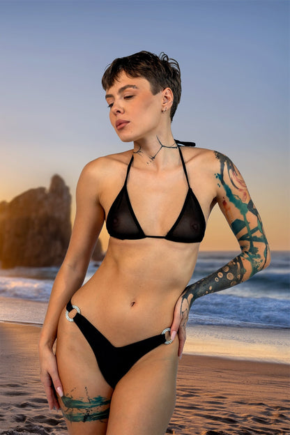 A woman models the V-Bottom Thong & Rio Bikini Bottoms by Brigitewear International, LLC on a sandy beach at sunset, with rocks and ocean in the background.