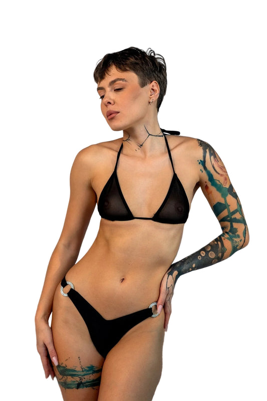 A woman models the V-Bottom Thong & Rio Bikini Bottoms by Brigitewear International, LLC on a sandy beach at sunset, with rocks and ocean in the background.