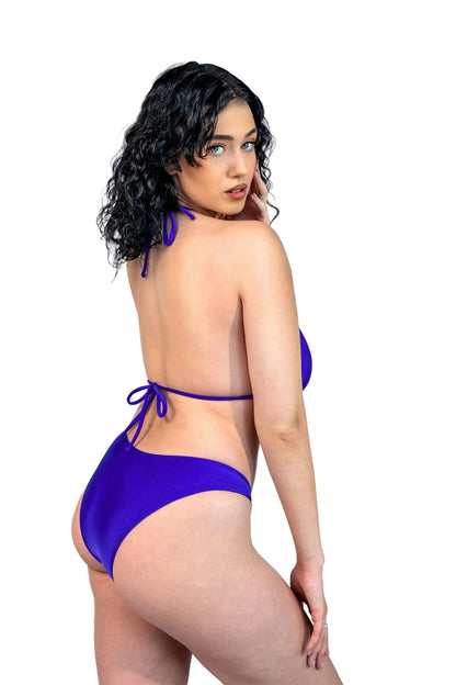 A woman with curly dark hair models the Brigitewear International V-Bottom Thong & Rio Bikini Bottoms in purple against a plain light gray background.